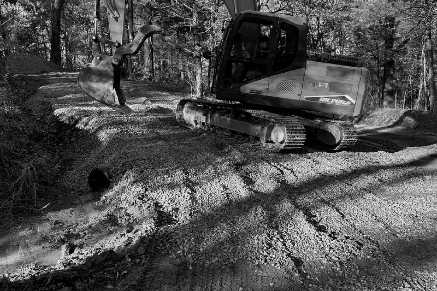 Excavator working gravel path with a drainage pipe exposed at the roadside