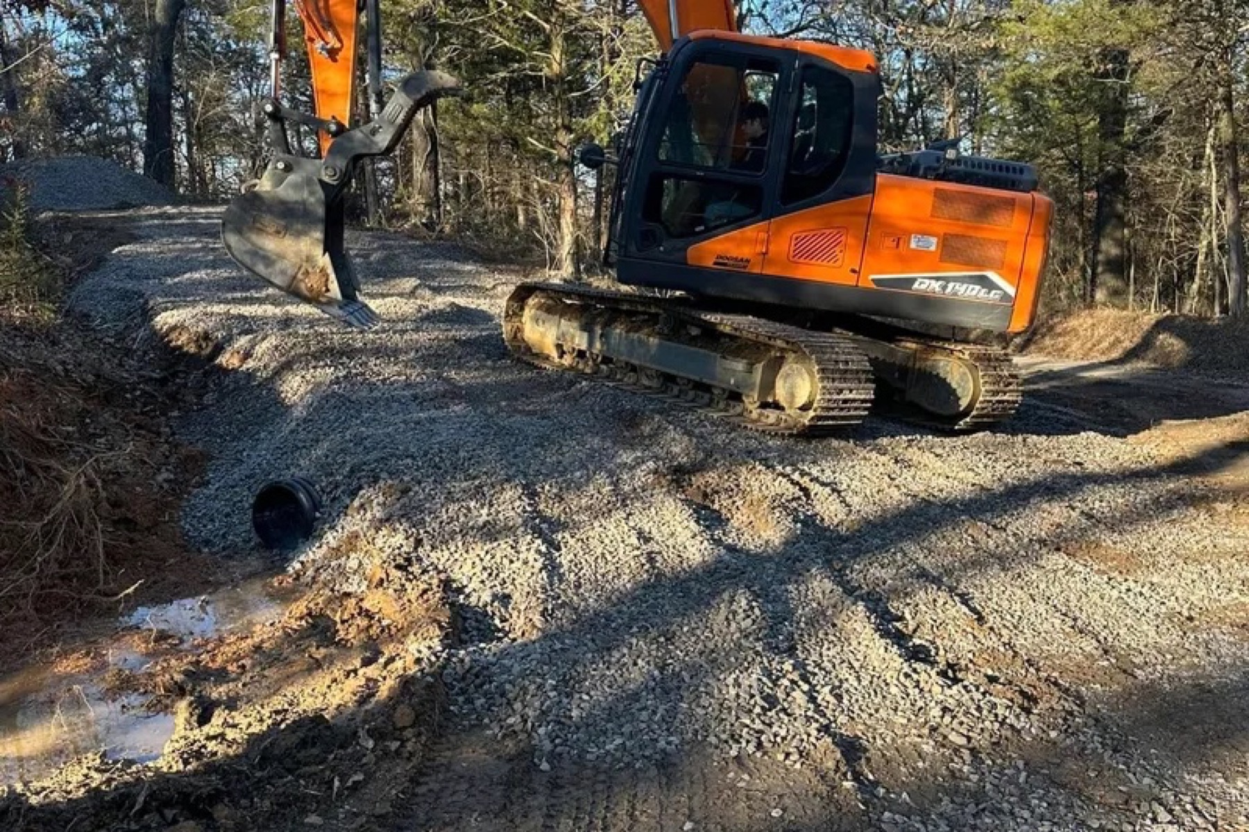 Excavator working gravel path with a drainage pipe exposed at the roadside