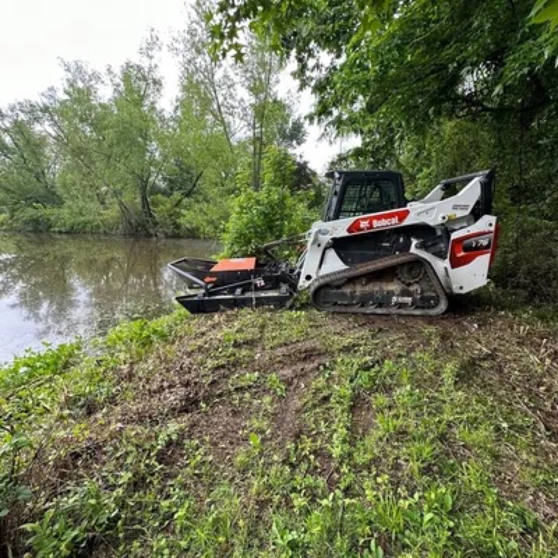 Bobcat with mower attachment clearing brush along a wooded shoreline